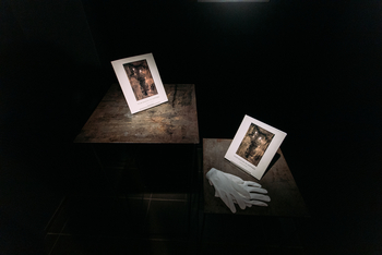 Zwei Fotografien stehen auf zwei nebeneinanderstehenden Holztischen ein paar weiße Handschuhe liegt daneben