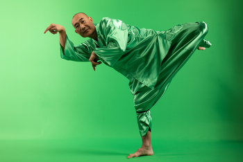 Ein Mann trägt einen grünen Stoffeinteiler steht auf einem Bein und steht vor einem grünen Hintergrund