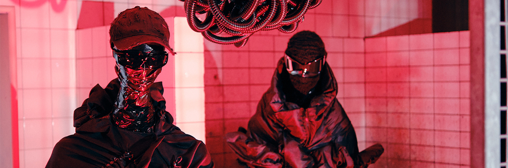 Zwei Aliens stehen in einem rot beleuchteten Raum und tragen Sonnenbrillen und schwarze Mäntel