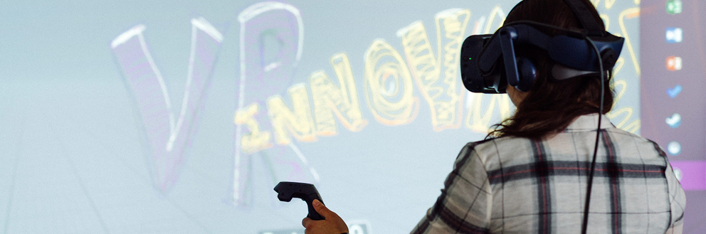 Eine Studentin hat eine Virtual Reality-Brille auf und steht vor einer Beamerfläche..